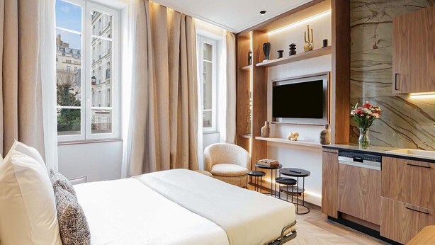 A hotel room with a bed, a chair, a TV, a small table, and a built-in kitchen area.