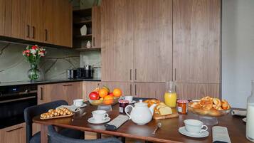 Une table de cuisine moderne, en bois, est dressée pour le petit-déjeuner : on y trouve un panier de fruits, des viennoiseries et une théière.