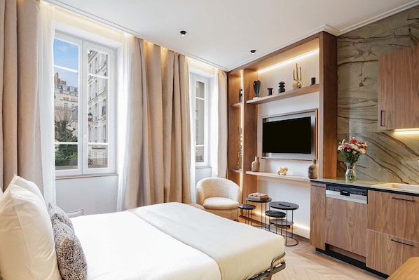 Habitación de hotel con cama, zona de estar con una silla y mesitas, cocina integrada con televisor y vistas de la ciudad a través de amplias ventanas.