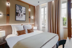 Habitación de hotel con una cama grande, cabecera con luces empotradas, un cuadro enmarcado sobre la cama y una ventana con cortinas.