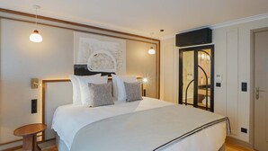 A modern hotel room with a large bed, a small side table, a painting above the bed, and a bathroom visible through an open door.