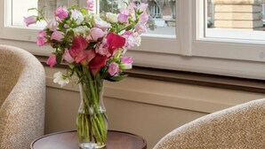 A window with a view of a bridge and buildings, a vase with flowers on a table, and a chair.