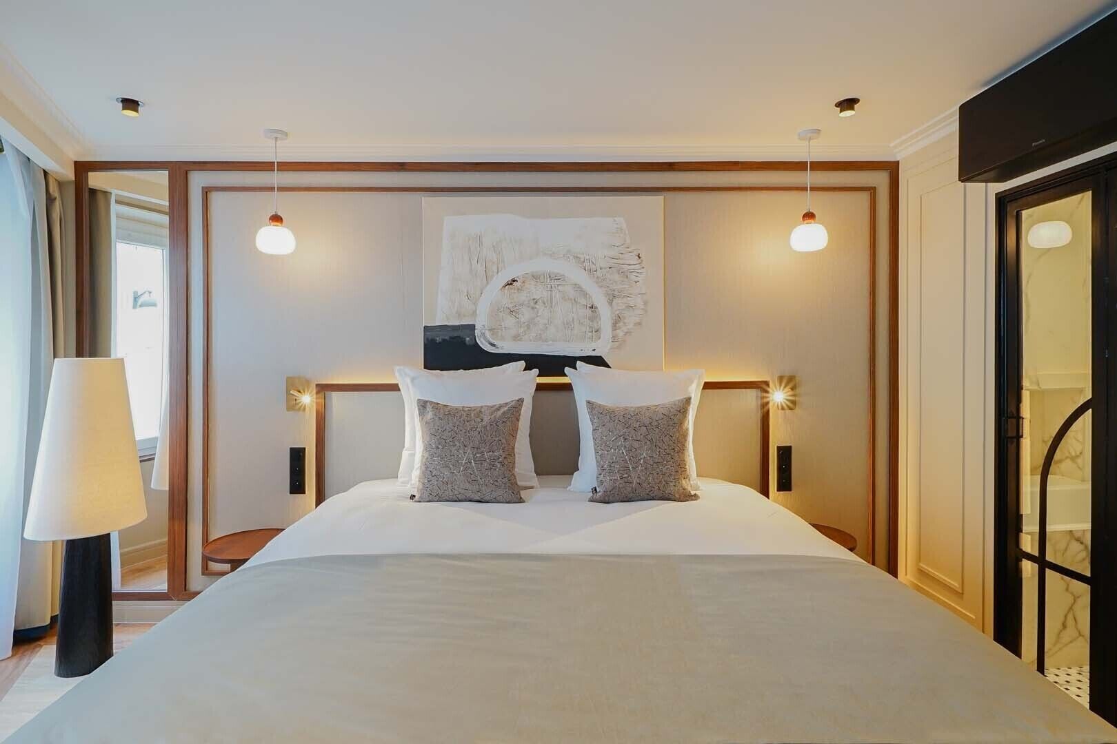 Een moderne hotelkamer met een groot bed, twee bedlampen, een groot abstract schilderij boven het bed en uitzicht op de stad door het raam.