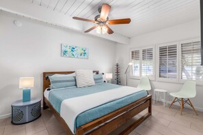 Une chambre à coucher avec un lit en bois, un ventilateur de plafond, une table de chevet avec une lampe, une chaise et une fenêtre avec des stores.
