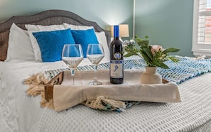 A neatly made bed with a headboard, two pillows, a bottle of wine, two glasses, and a small potted plant on a tray.