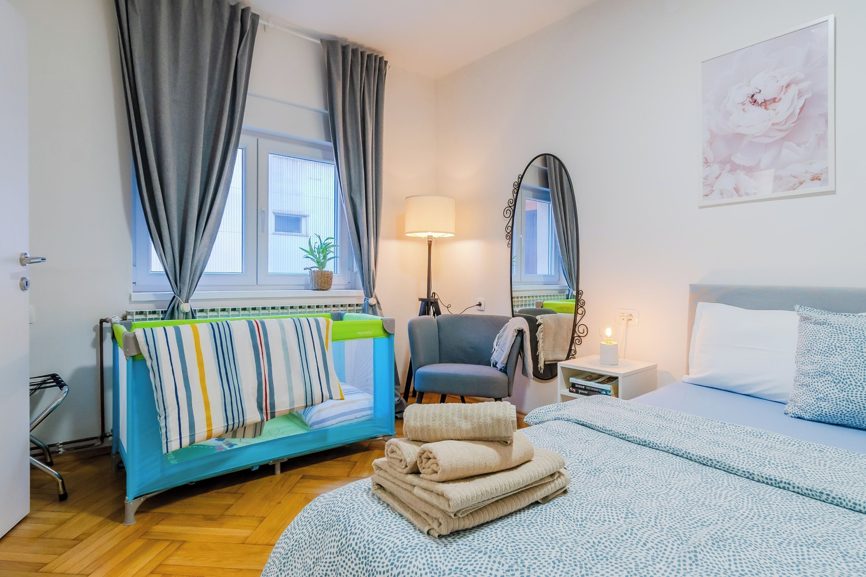 A hotel room with a bed, a chair, a bedside table with a lamp, a mirror, and a window with curtains.