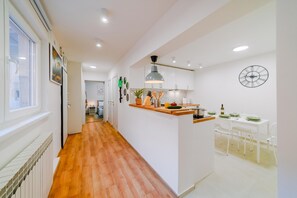 A modern kitchen with wooden flooring, white cabinets, a central island, and a dining area with white chairs. - Peaceful Nest In City Center (Zagreb)