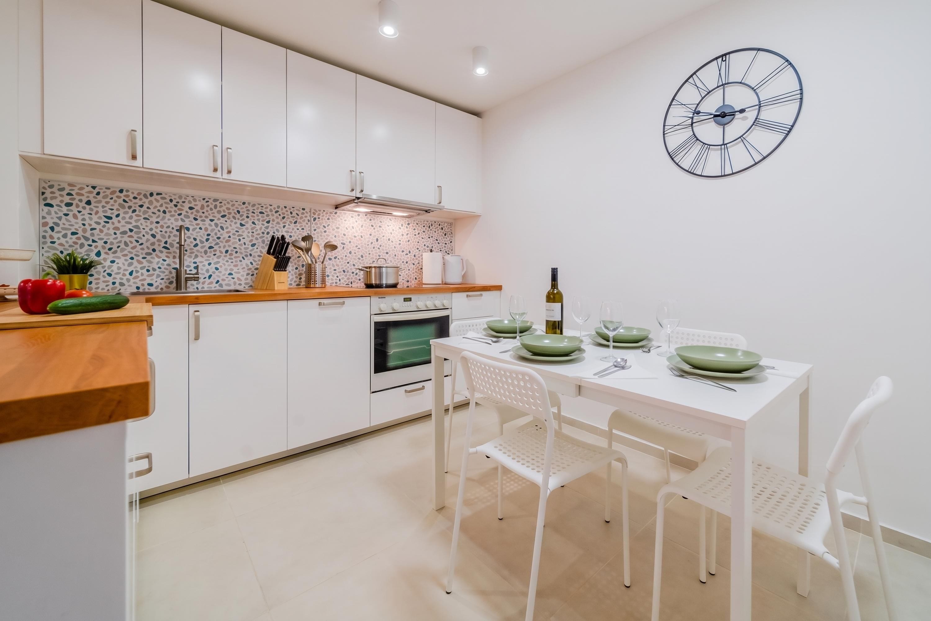 A modern kitchen with white cabinets, a countertop with kitchen utensils, a microwave, and a dining area with a table set for two.