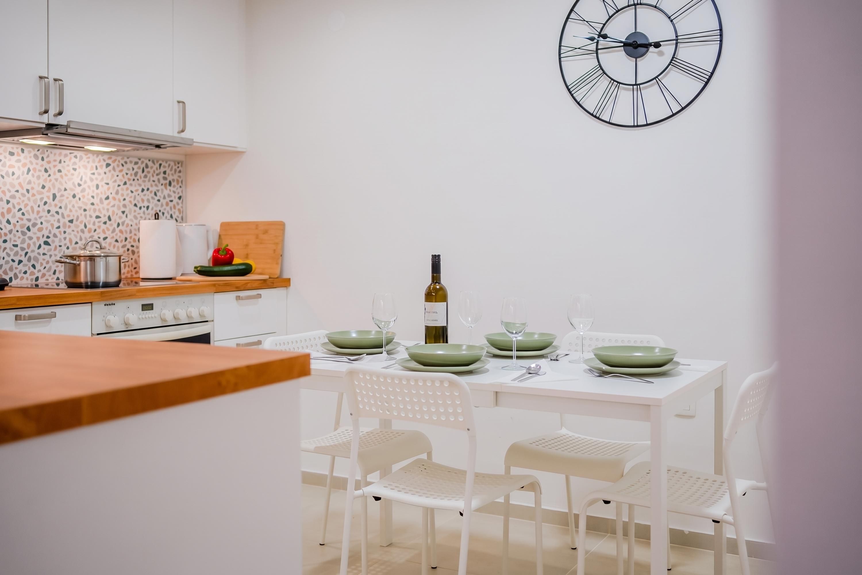 Une cuisine moderne avec des meubles blancs, un plan de travail en bois et une table à manger dressée avec de l’assiette verte et des verres à vin.