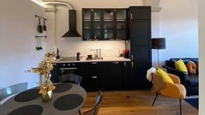 Eine moderne Küche mit dunklen Schränken und Holzbalkendecke, ein Essbereich mit rundem Tisch und Stühlen, sowie ein Wohnbereich mit Sofa und Lampe.
