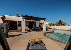 A poolside area with a covered seating area, two drinks on a table, and a clear blue sky.