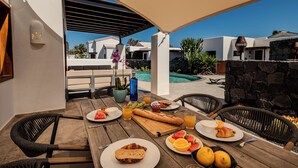 Una mesa de madera con alimentos para el desayuno, sillas exteriores y vistas a una piscina y edificios modernos.