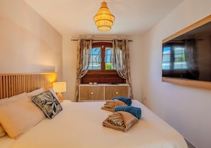 A hotel room with a bed, a bedside lamp, a wooden headboard, a window with curtains, and a television mounted on the wall.