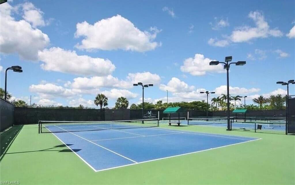 An outdoor tennis court with multiple courts, surrounded by a fence, and equipped with overhead lighting.