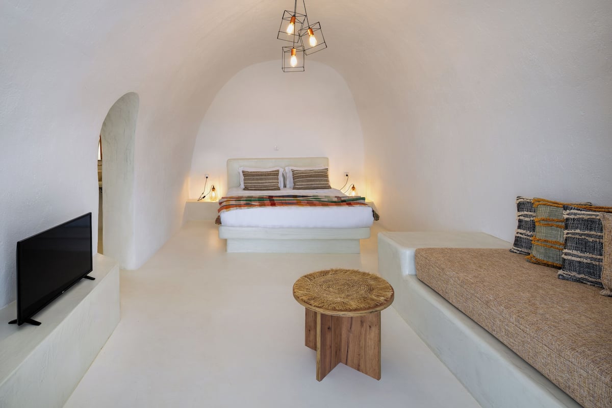 A minimalist bedroom with a bed, a TV, a small wooden stool, and a sofa.