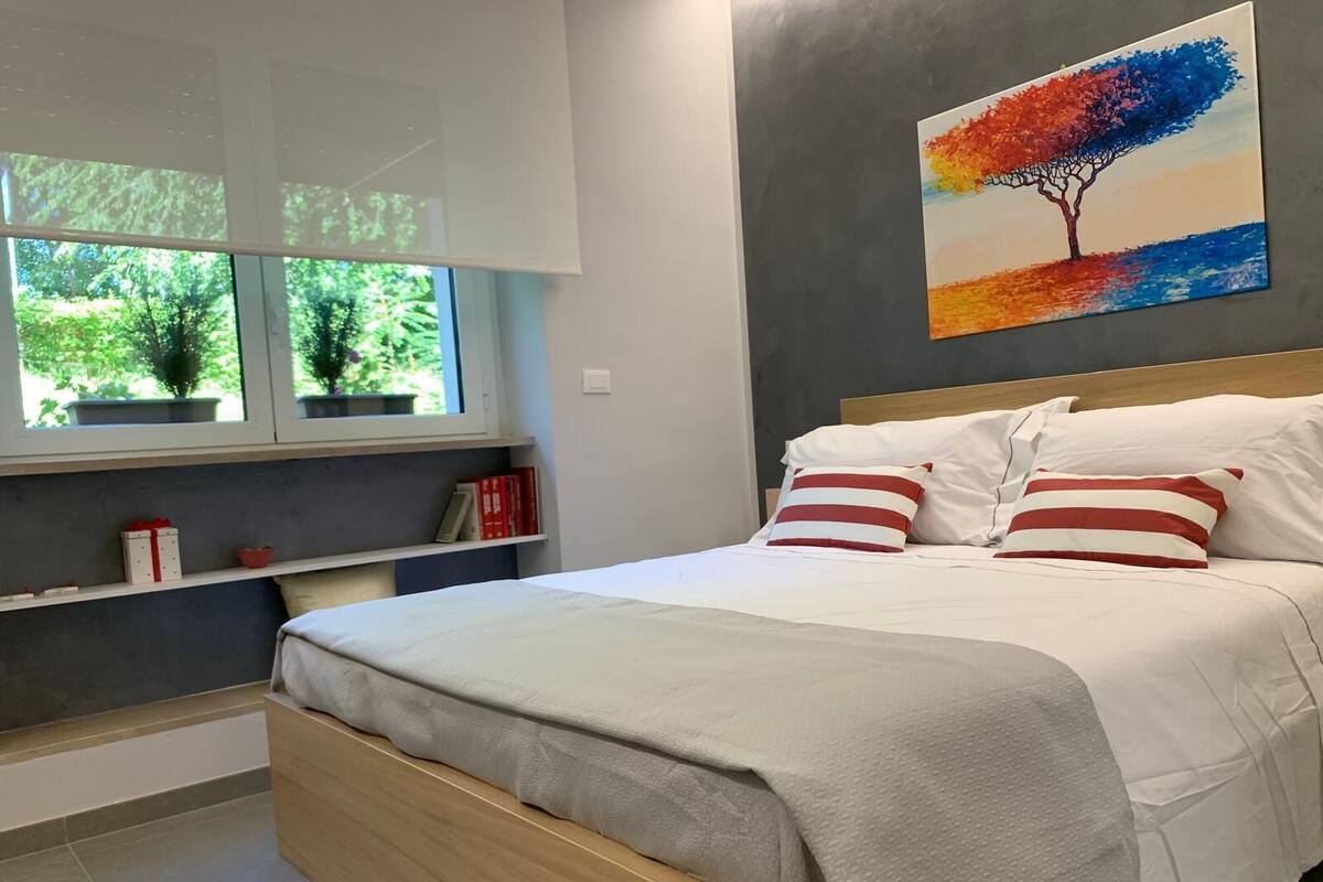 Egy modern hálószoba, amelyben található egy ágy, egy ablak, melyből zöld tájra nyílik kilátás, egy könyvespolc és egy kép a falon.