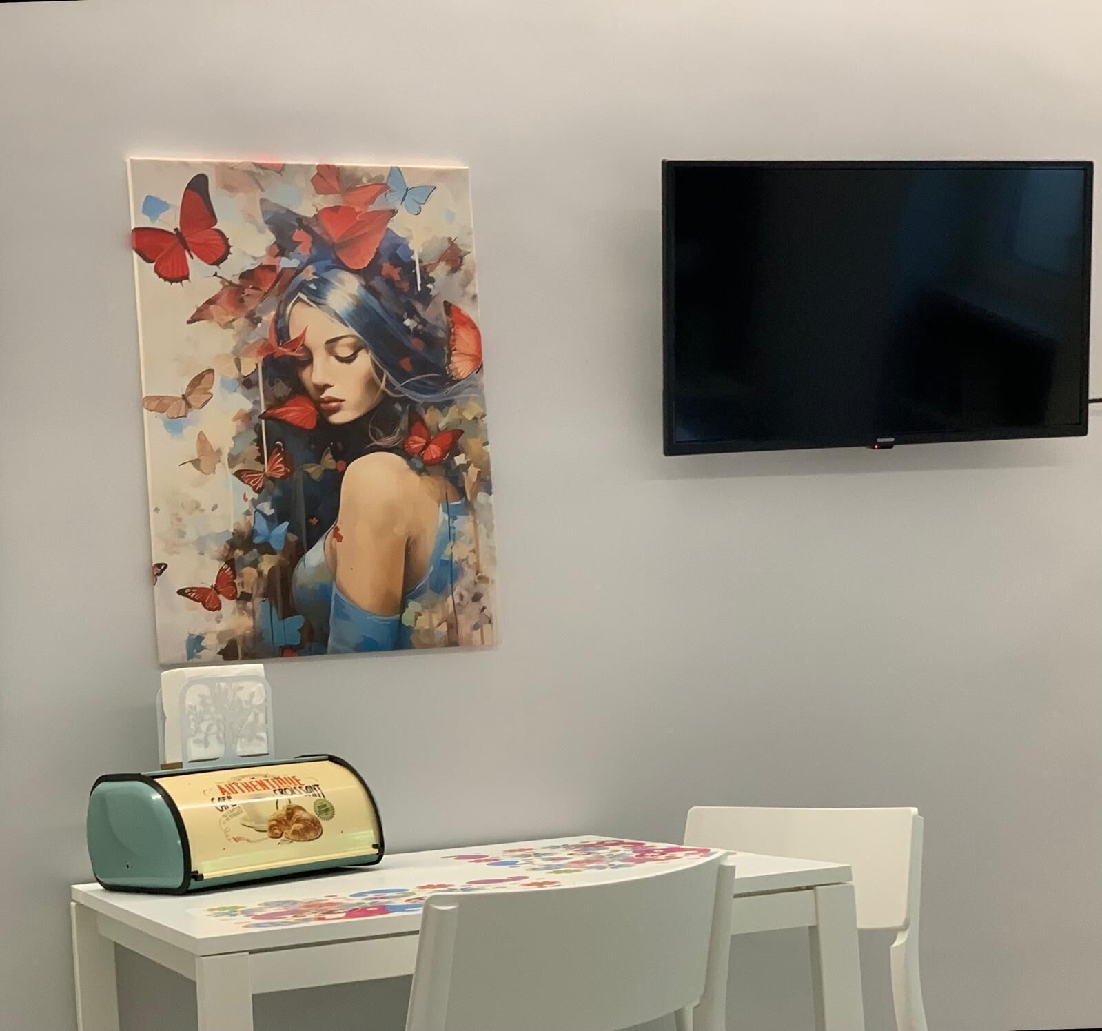A room with a flat-screen TV mounted on the wall, a table with chairs, and a painting with butterflies and a woman.