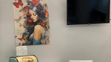 A room with a flat-screen TV mounted on the wall, a table with chairs, and a painting with butterflies and a woman.