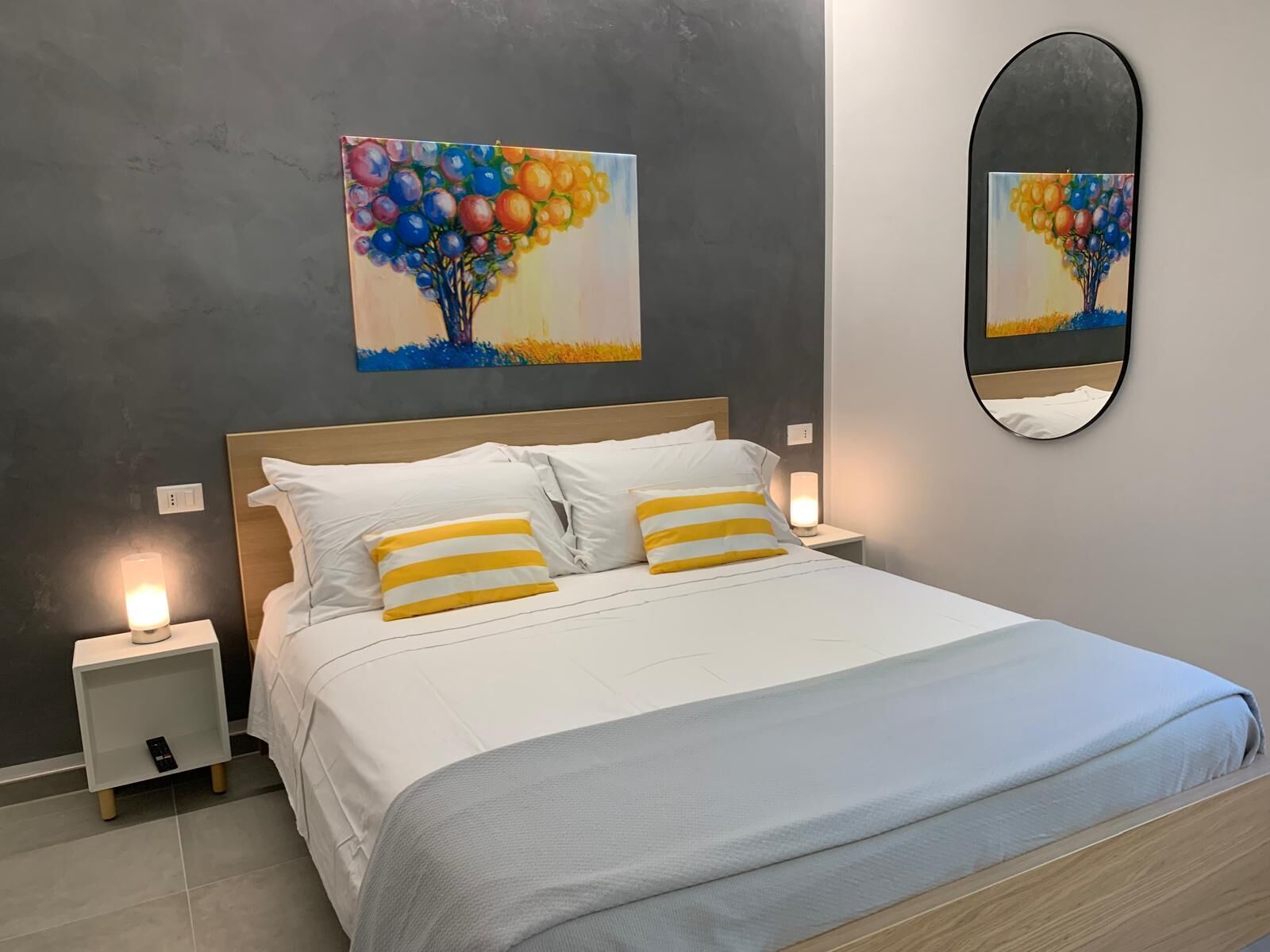 A bedroom with a bed, two bedside tables, a nightstand, a mirror, and a painting of a tree with balloons.