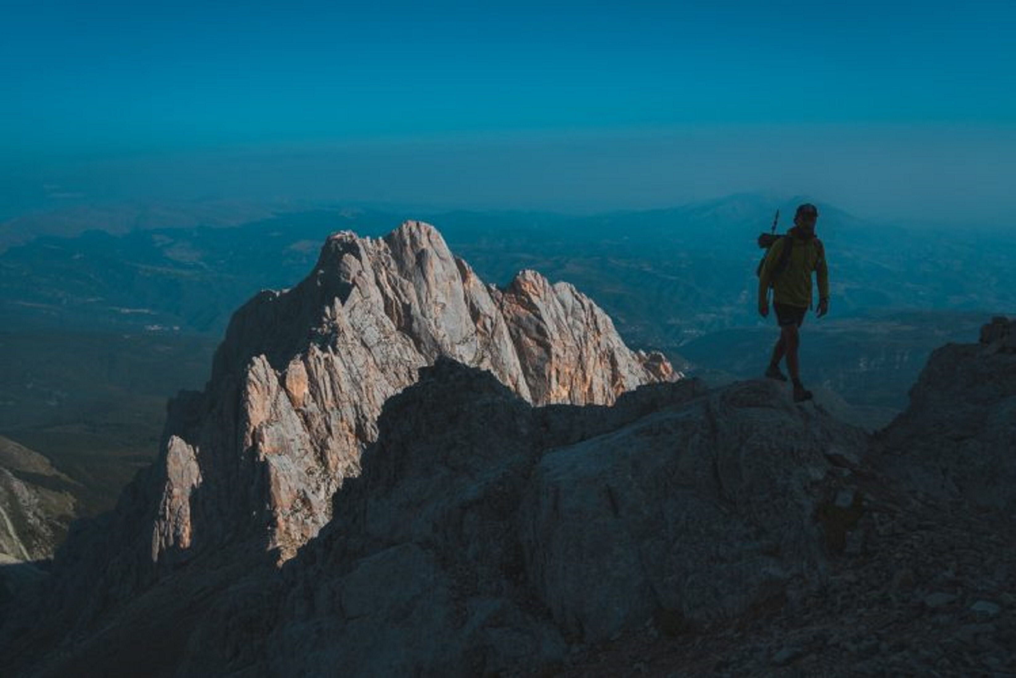 A person standing on a rocky peak with a backpack, overlooking a mountainous landscape.