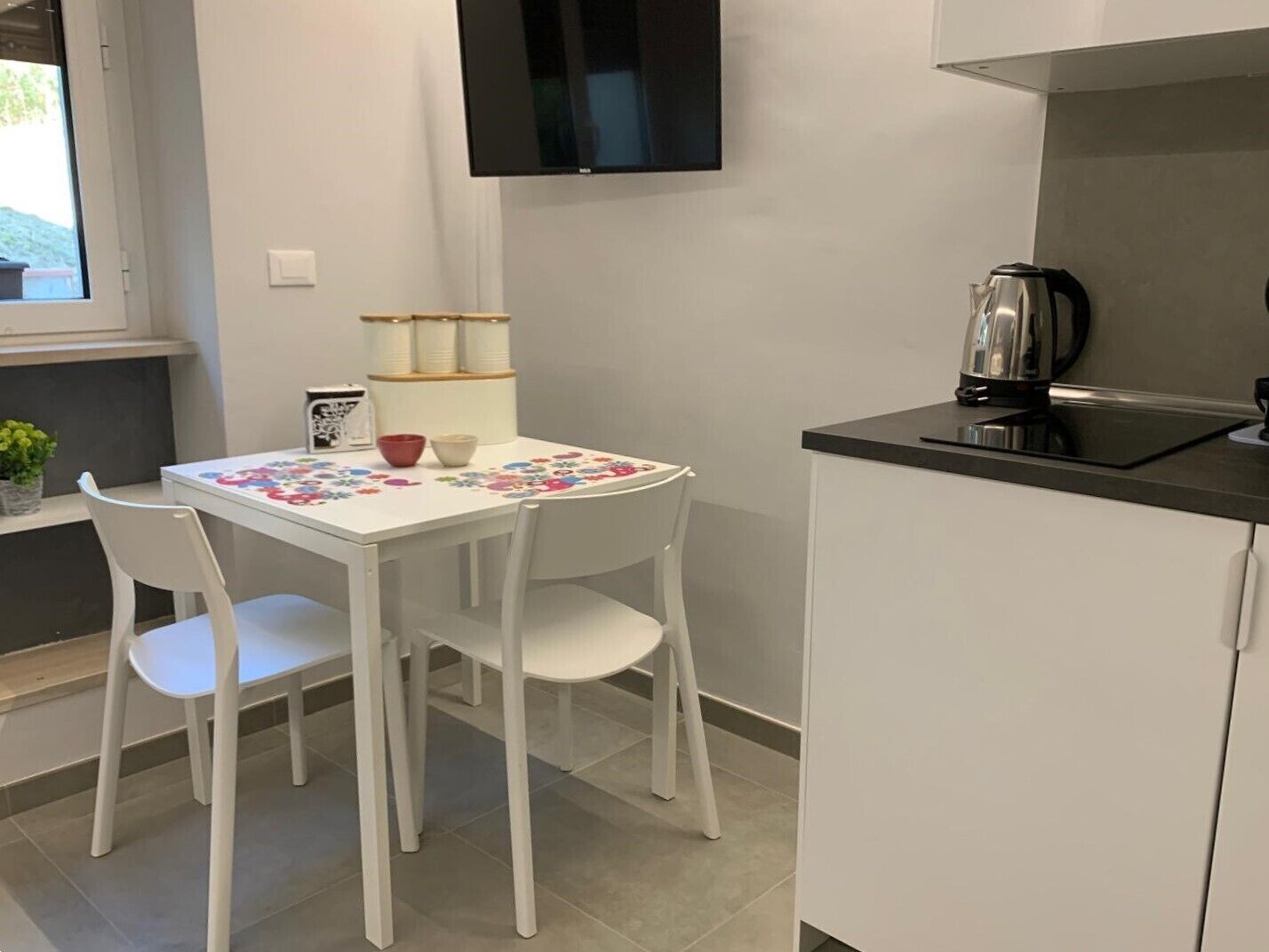Una cocina pequeña con una mesa blanca, dos sillas blancas, una encimera negra y una tetera.