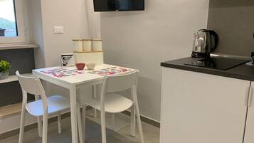 Una cocina pequeña con una mesa blanca, dos sillas blancas, una encimera negra y una tetera.