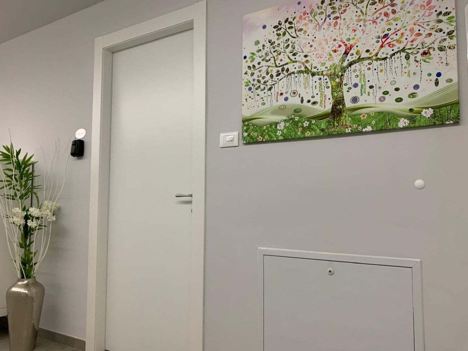 Un couloir avec une porte blanche, un luminaire fixé au mur, un tableau décoratif représentant un arbre et une plante en pot.