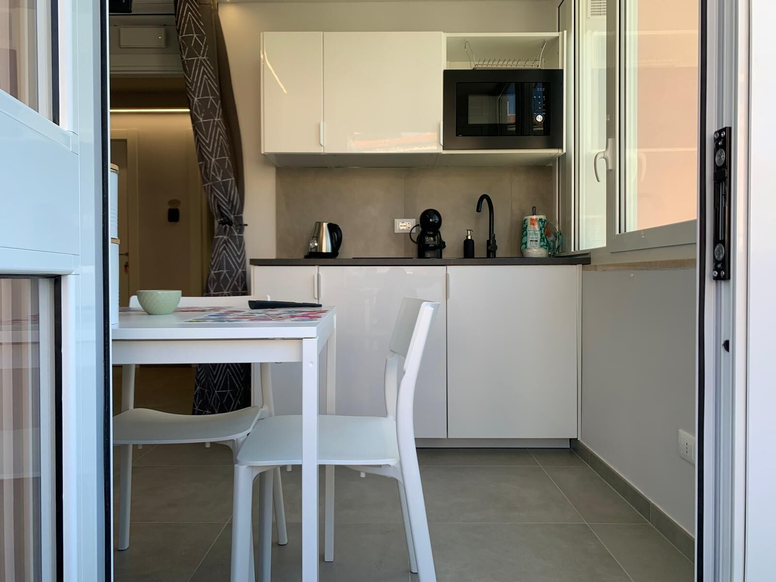 A compact kitchen with white cabinets, a microwave, a coffee maker, and a kettle. There is a small dining table with two chairs.