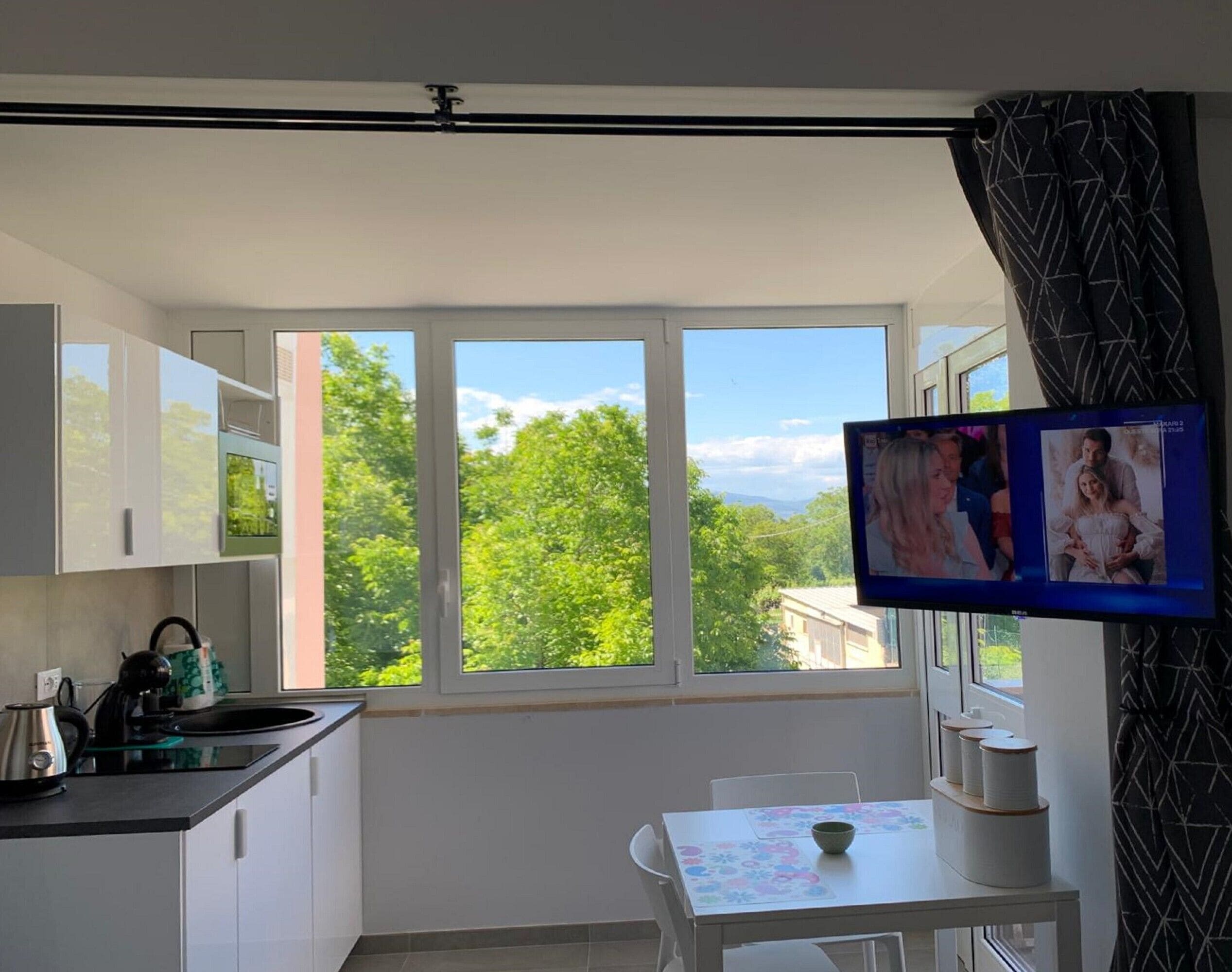 A modern kitchen with a large window, a dining area with a table, and a TV mounted on the wall.