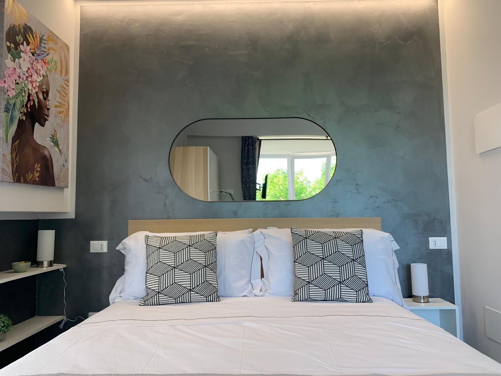 Un dormitorio moderno con una cama grande, almohadas estampadas con motivos geométricos, un espejo redondo y un cuadro en la pared.