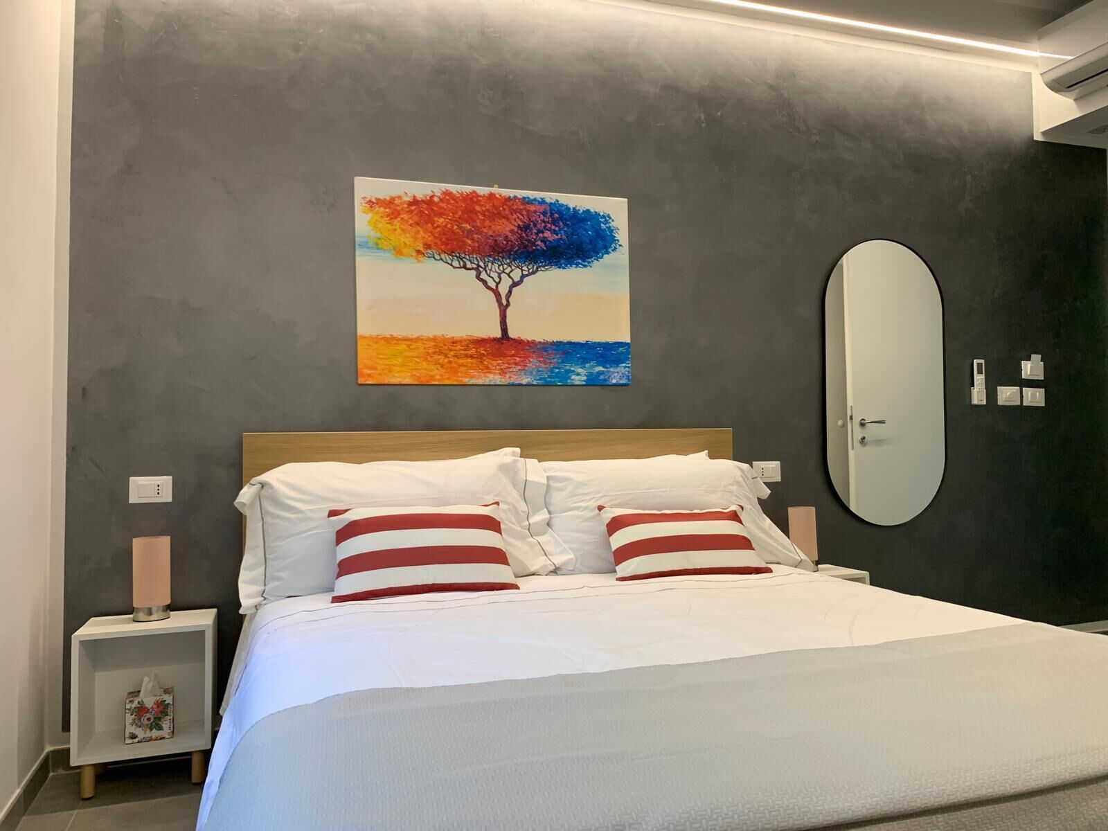 Quarto moderno com cama, mesa de cabeceira, espelho e pintura de uma árvore.