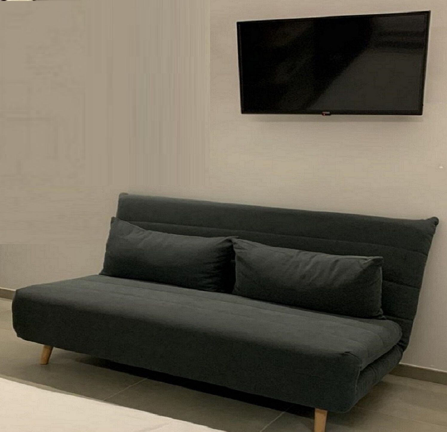 A dark grey sofa with wooden legs against a light-colored wall, with a flat-screen TV mounted above it.
