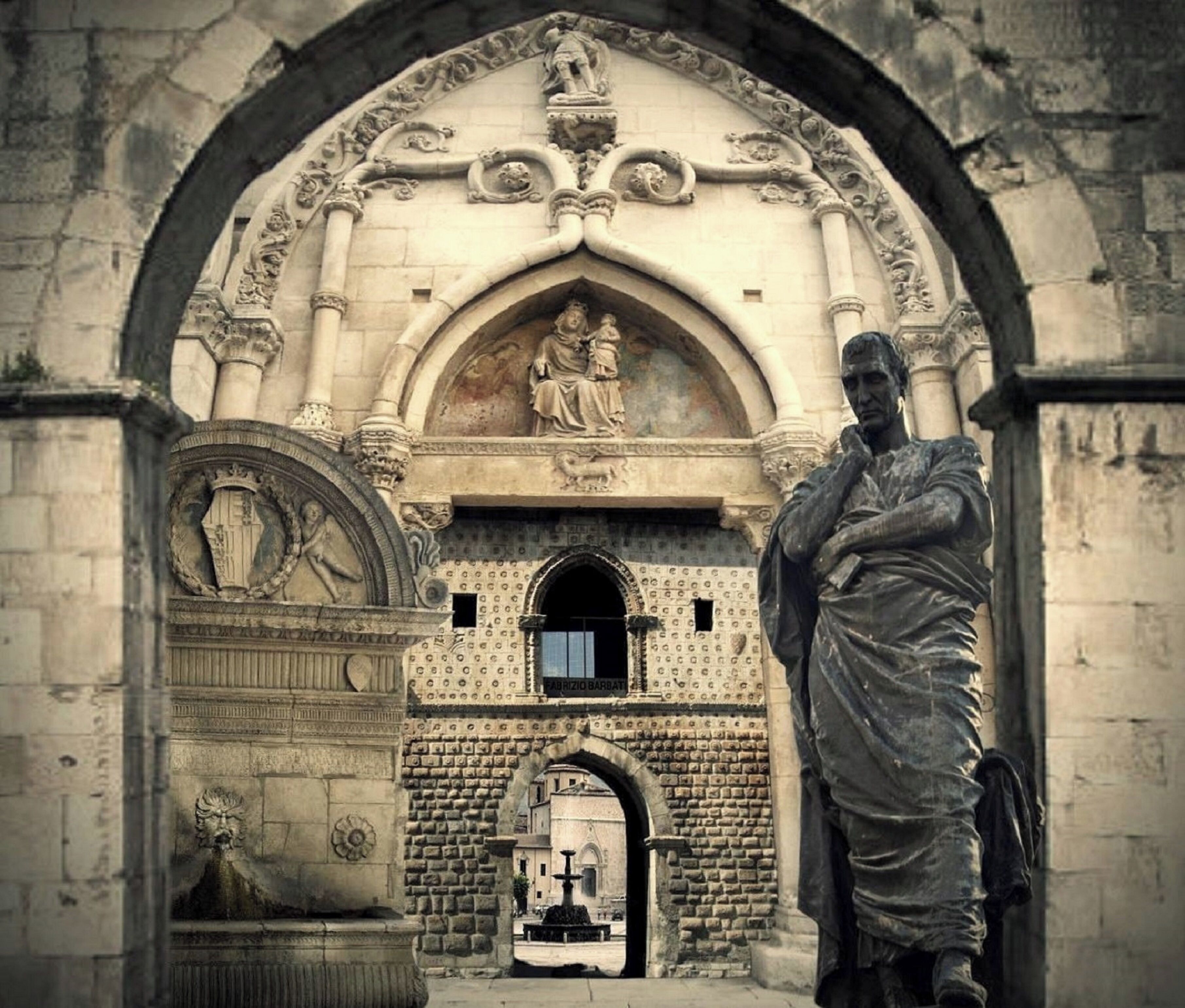 Ein Eingang eines historischen Gebäudes mit aufwendigen Steinmetzarbeiten, einer Statue und einem Brunnen.