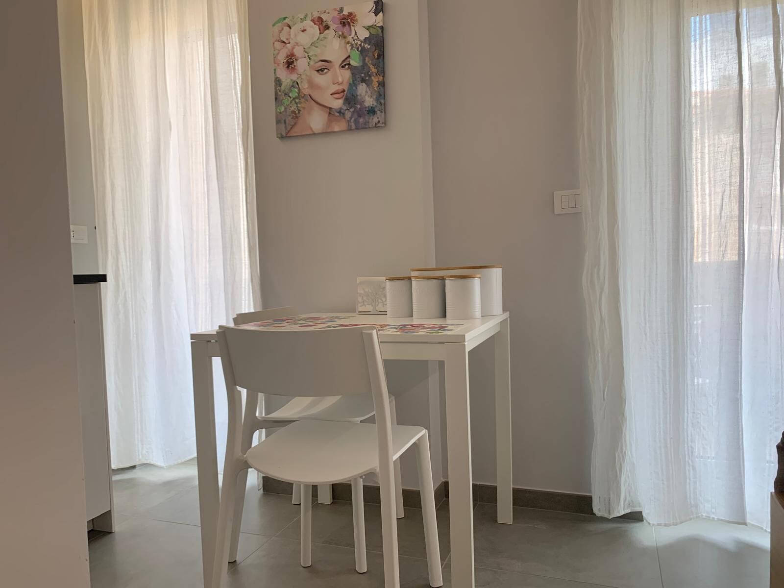 A dining area with a white table and chairs, a painting on the wall, and sheer curtains.