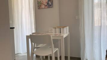 A dining area with a white table and chairs, a painting on the wall, and sheer curtains.