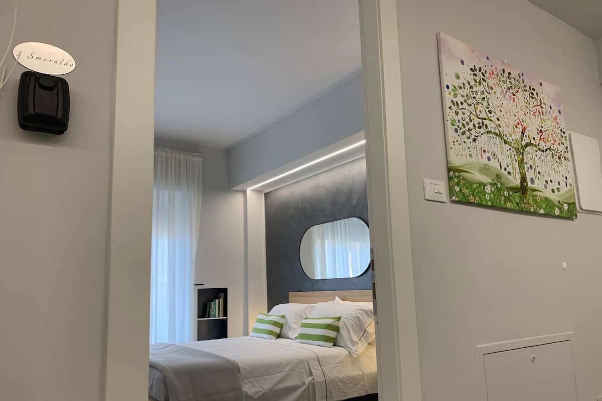 Egy modern hálószoba, amelyben található egy ágy, egy éjjeliszemélyzet, egy tükör, egy falra szerelt lámpa és egy festmény.