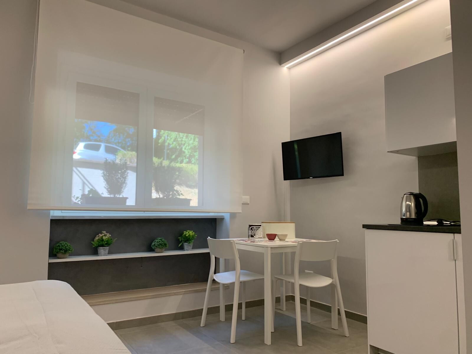 Ein kleiner, moderner Raum mit einem Bett, einem Fenster mit Blick auf ein Auto und Grünflächen, einem Tisch mit Stühlen, einem Flachbildfernseher und einem Wasserkocher auf der Ablagefläche.