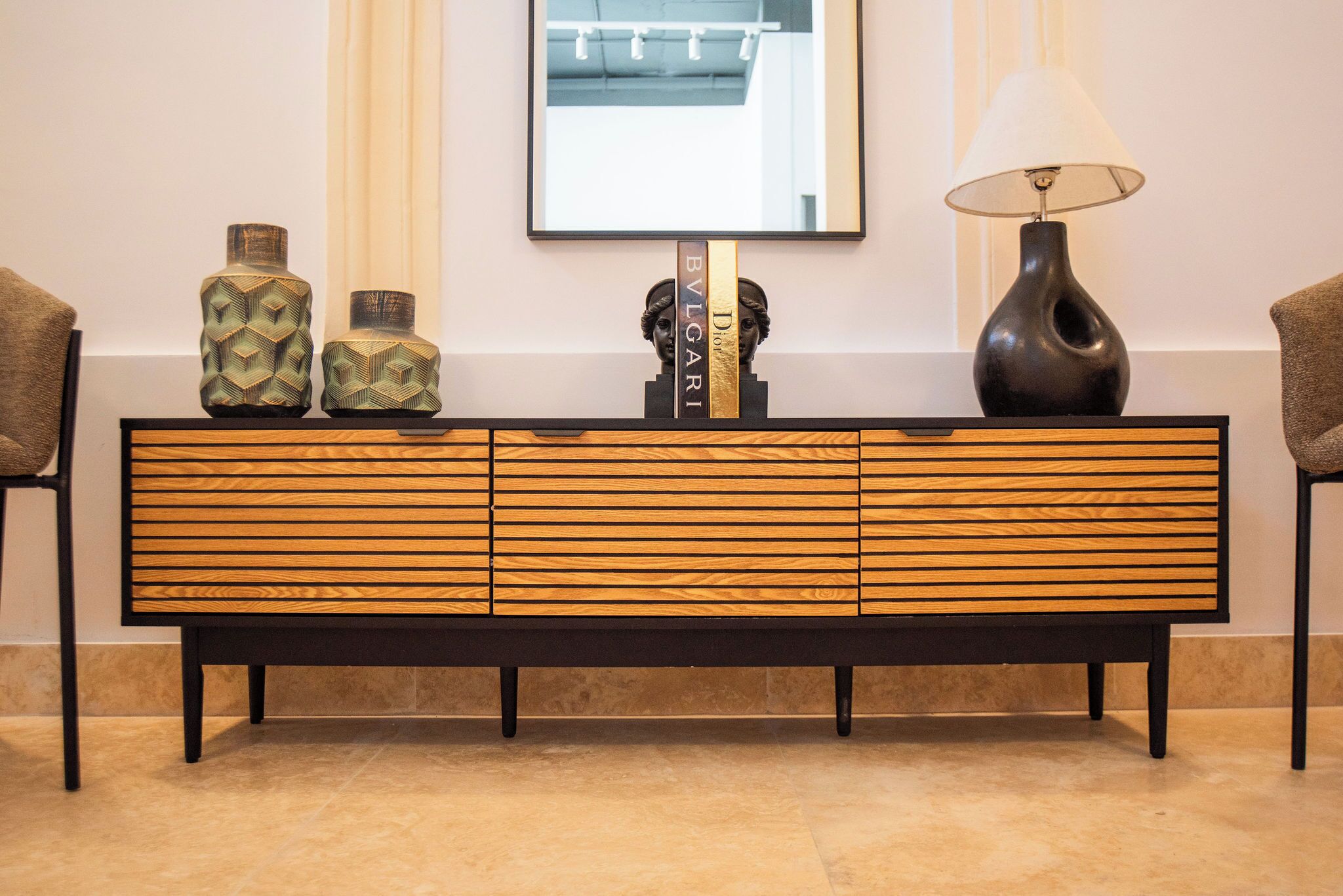 A modern, wooden console table with three decorative vases and a lamp, flanked by two chairs.