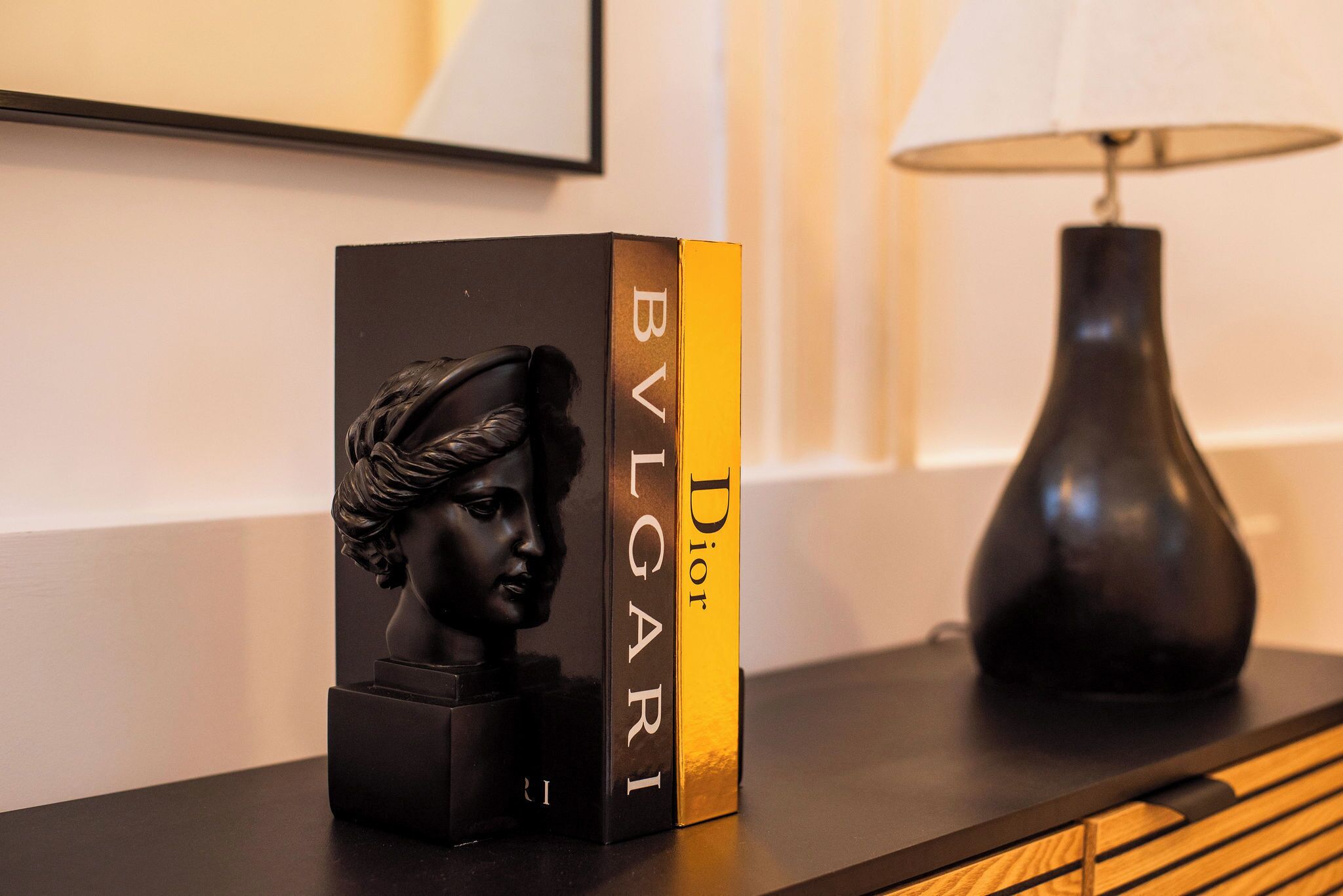 Musta rintakuva on sijoitettu mustan laatikon päälle, ja laatikossa on merkintä ”BVLGARI”. Sen vieressä on musta lamppu, ja kaikki on asetettu puupinnan päälle.