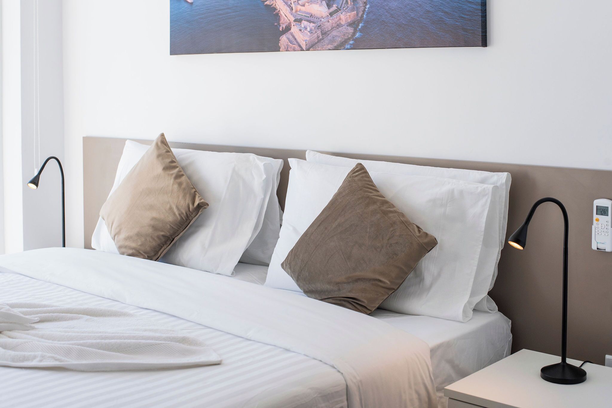 Una cama bien hecha con ropa de cama blanca y beige, una obra de arte colgada en la pared y una mesita de noche con una lámpara.