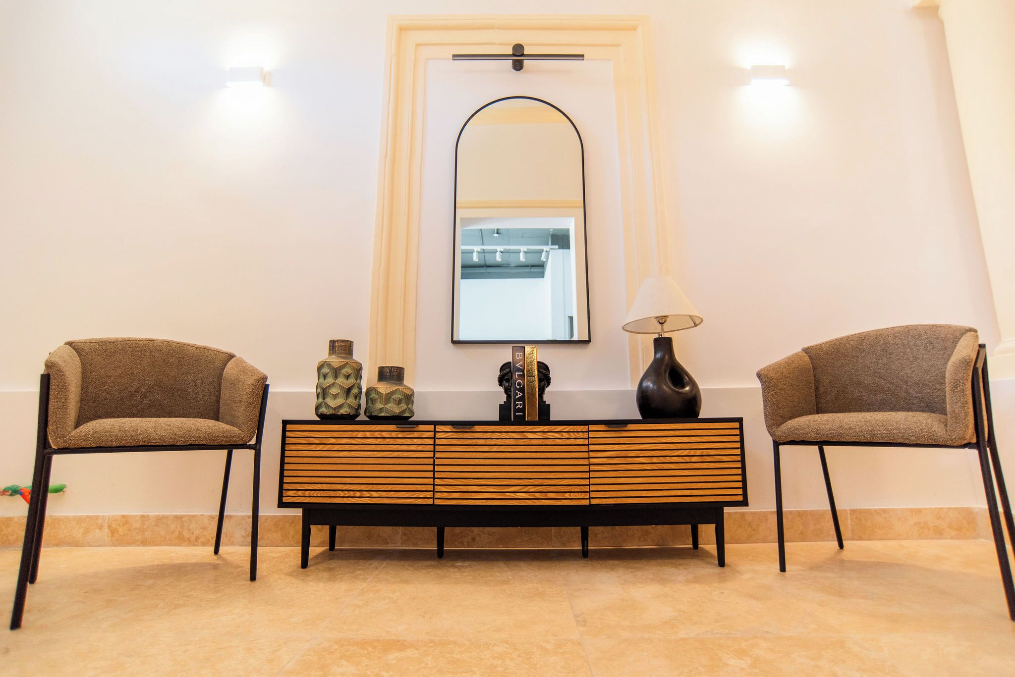 A room with a mirrored wall, two chairs, a wooden console table with decorative items, and a lamp.