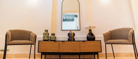 A room with a mirrored wall, two chairs, a wooden console table with decorative items, and a lamp.