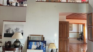 Um loft espaçoso com pé-direito alto, vigas de madeira e um grande lustre. Possui vários níveis, incluindo um mezanino, uma escada e vista para o céu.