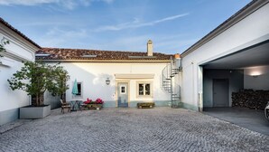 Um pátio com piso de lajes, um edifício branco com telhado em telhas, uma escadaria e um banco.