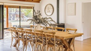 Un coin repas avec une table et des chaises en bois, une grande horloge au mur et une vue sur un terrain de jeux extérieur, visible à travers de grandes fenêtres.