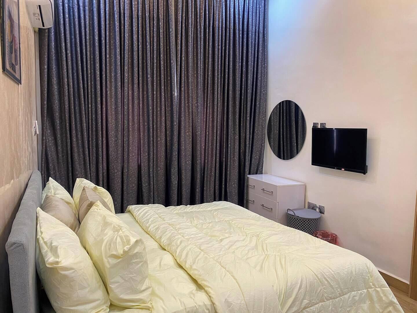 Un dormitorio con cama, cortinas grises, televisor y un pequeño armario blanco.