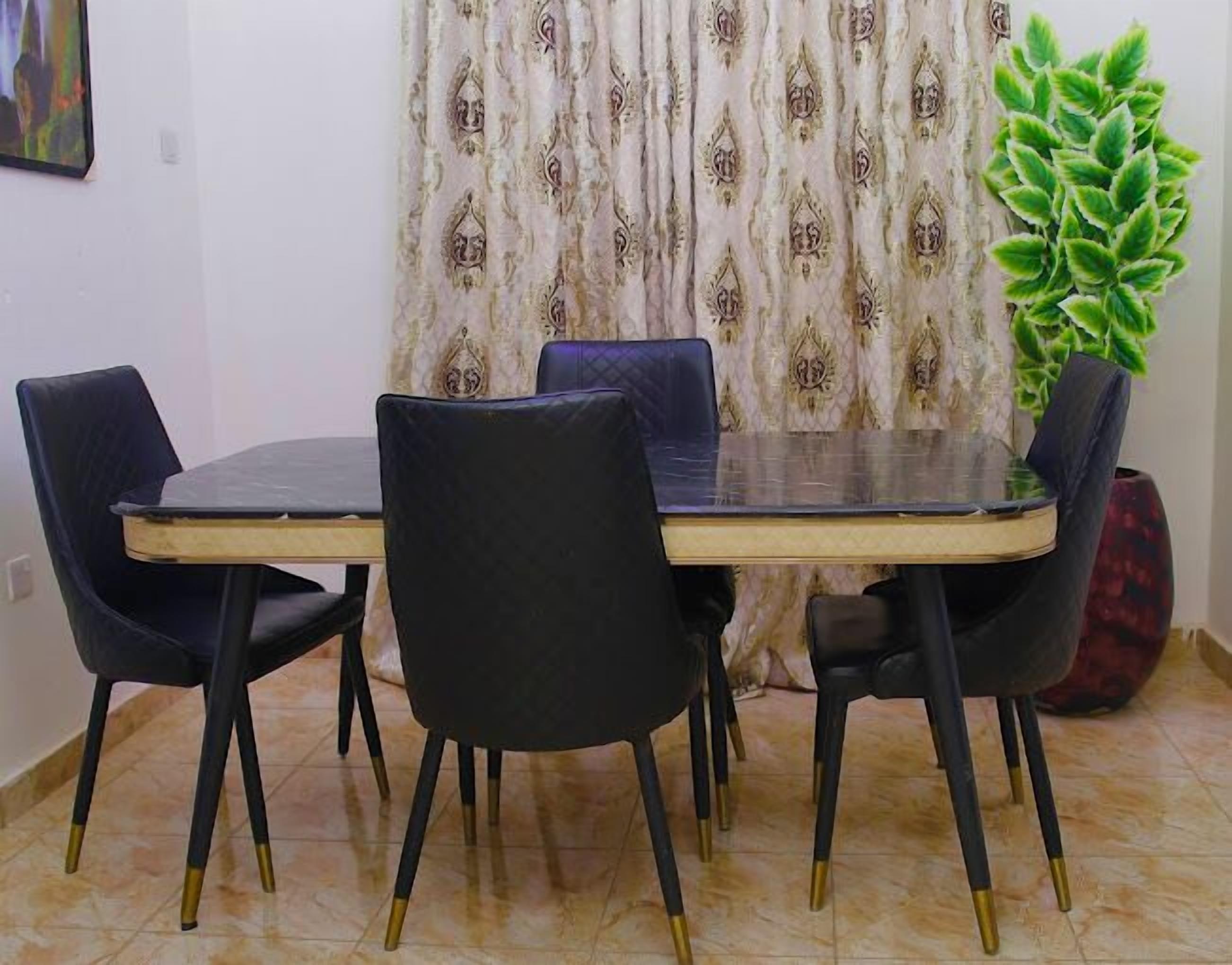Un coin repas comprenant une table rectangulaire, quatre chaises noires aux pieds dorés, un rideau à motifs et une plante verte.