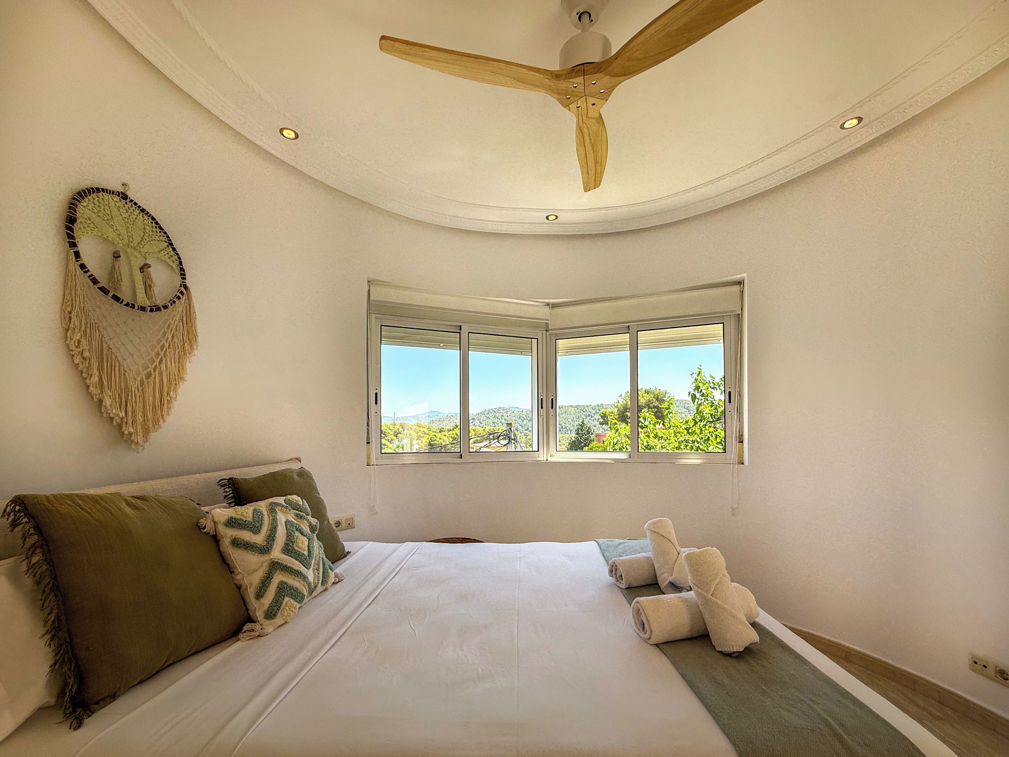 Ein Schlafzimmer mit Bett, Kissen, Wandbehang aus Makramee, Deckenventilator und einem Fenster mit Blick.