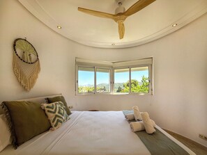 A bedroom with a bed, pillows, a macrame wall hanging, a ceiling fan, and a window with a view. - Villa Claudia by Hello Homes Sitges (Sitges)