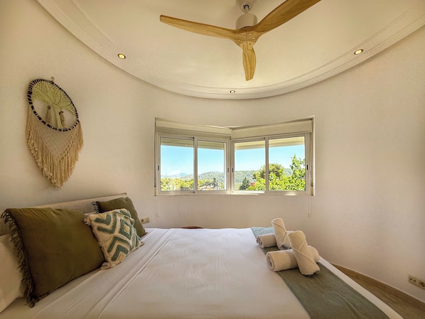 A bedroom with a bed, pillows, a macrame wall hanging, a ceiling fan, and a window with a view. - Villa Claudia by Hello Homes Sitges (Sitges)